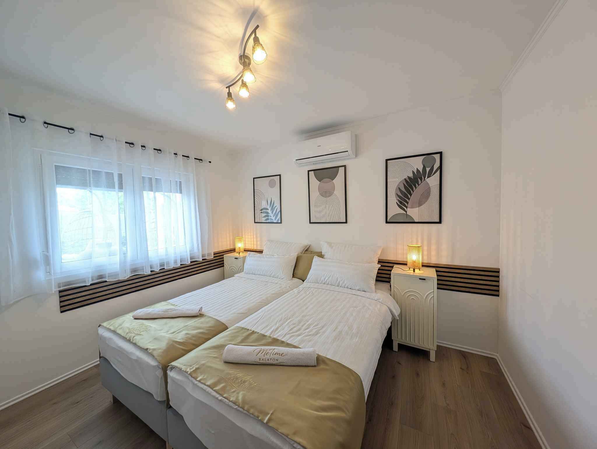 A double bed with a beige bedspread, two bedside tables with lamps, a window with sheer curtains, and wall art.