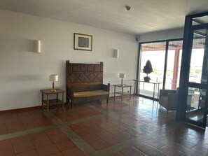 Quarto com um banco de madeira, duas mesinhas com luminárias, um quadro pendurado na parede e uma janela grande com vista externa.