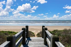 Ein hölzerner Steg führt zu einem Strand mit blauem Himmel und Blick auf das Meer.