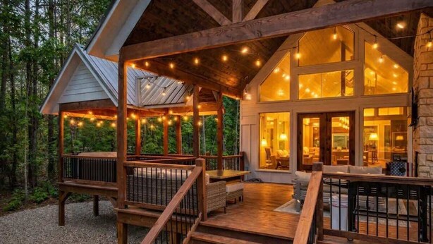 Una terraza de madera con mobiliario exterior, un espacio cubierto con luces colgantes y un edificio de amplios ventanales y fachada de piedra.