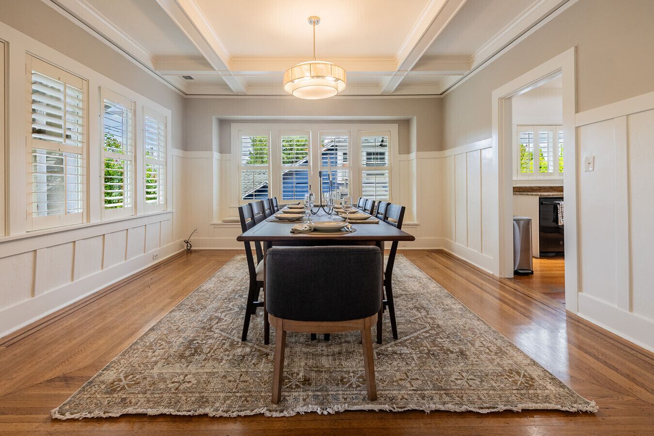 A dining room with a wooden table set for six, surrounded by chairs, with a patterned rug on the floor.