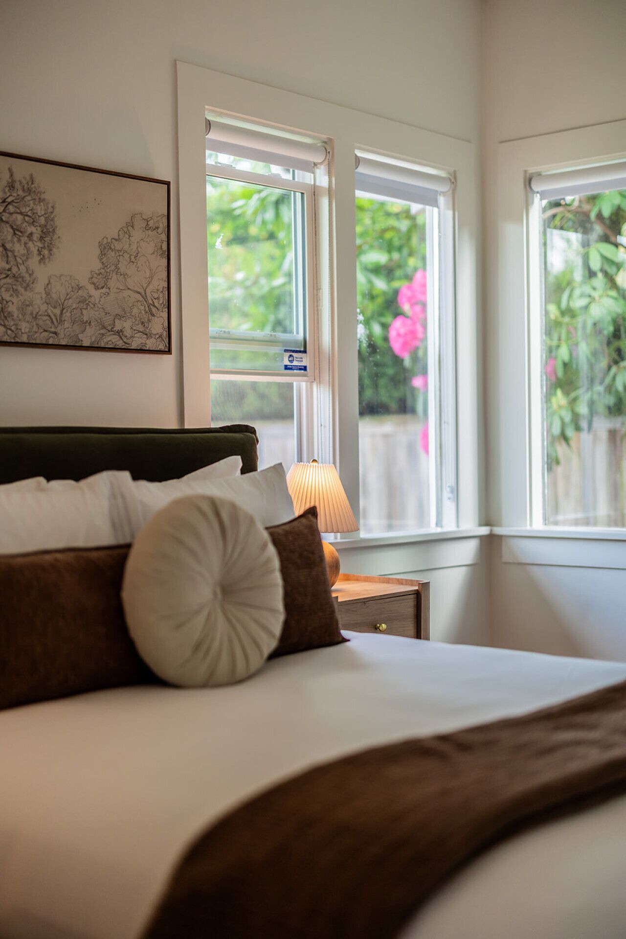 A bedroom with a bed, pillows, a nightstand, a lamp, and windows with a view of greenery.