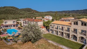 A multi-story hotel with a swimming pool, outdoor seating, and a view of the surrounding hills. - Grape Vine (Zakynthos)