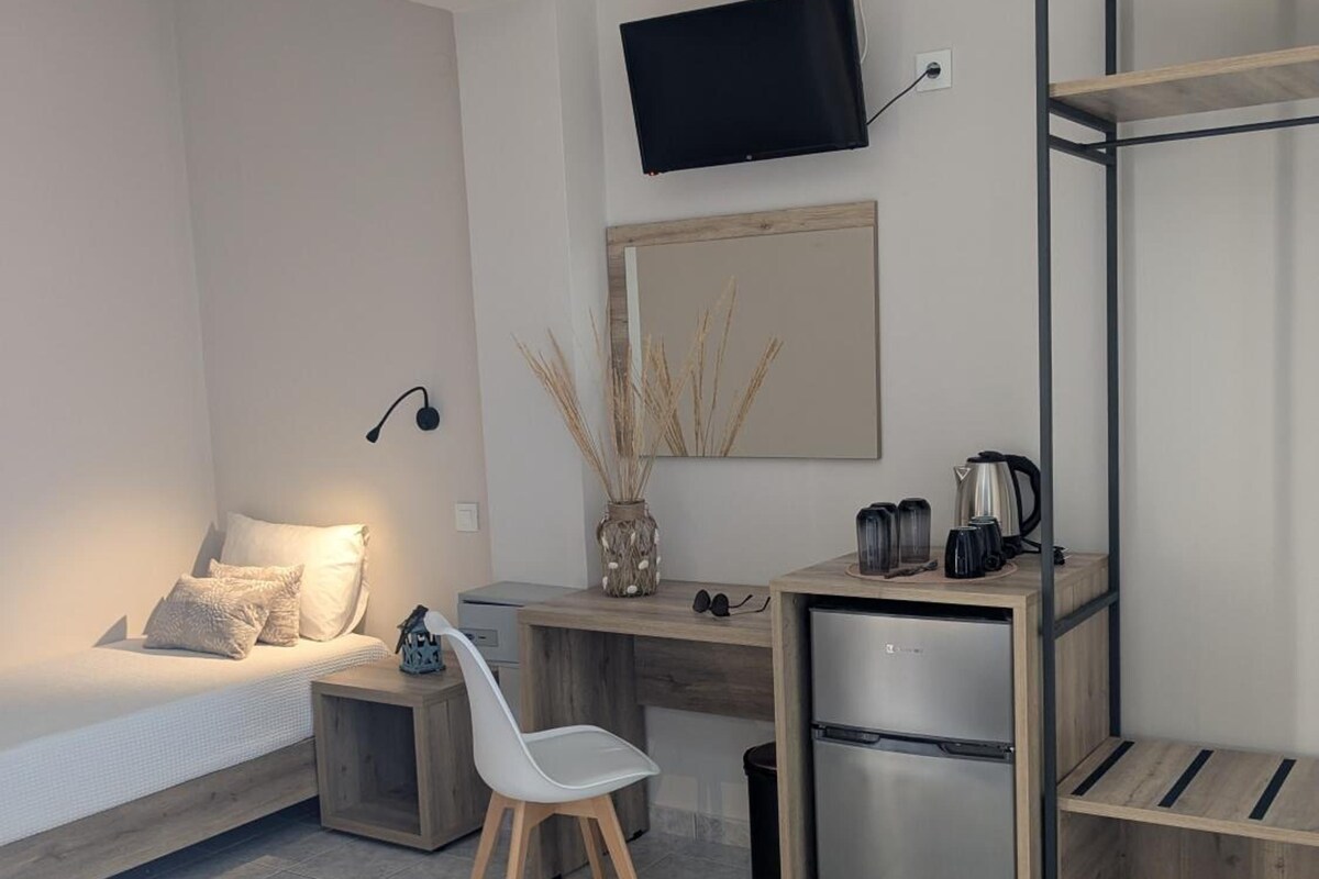 A compact bedroom with a bed, a desk, a chair, a TV, and a kitchenette.