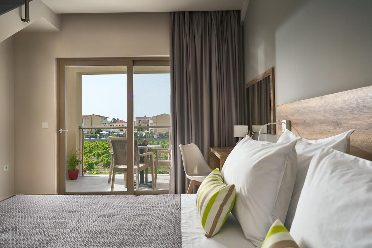 A hotel room with a bed, a chair, a table, and a view of the outside.