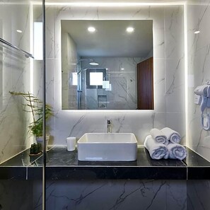 Un baño moderno con un espejo grande, un lavamanos rectangular, una planta en maceta y toallas enrolladas.