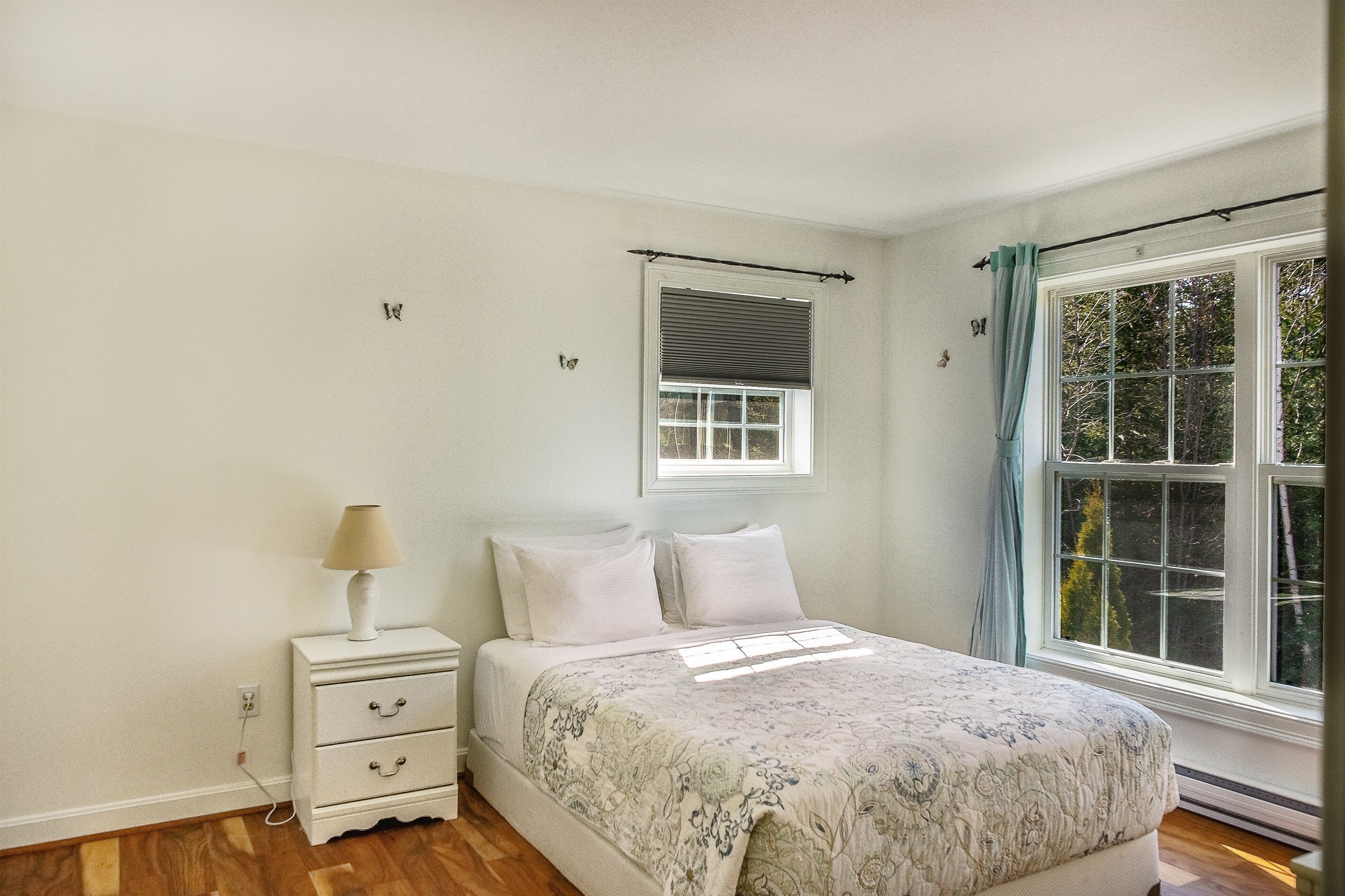 Une chambre à coucher comprenant un lit, une table de chevet, une lampe, une fenêtre avec des rideaux et un interrupteur fixé au mur.