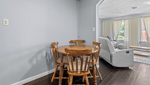 Een eetgedeelte met een ronde houten tafel, stoelen, een grijze bank en een raam met gordijnen.