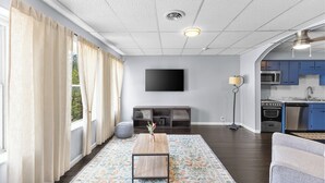 Een woonkamer met een flatscreen televisie, een bank, een salontafel, een vloerlamp en een keuken met blauwe kastjes en apparatuur.