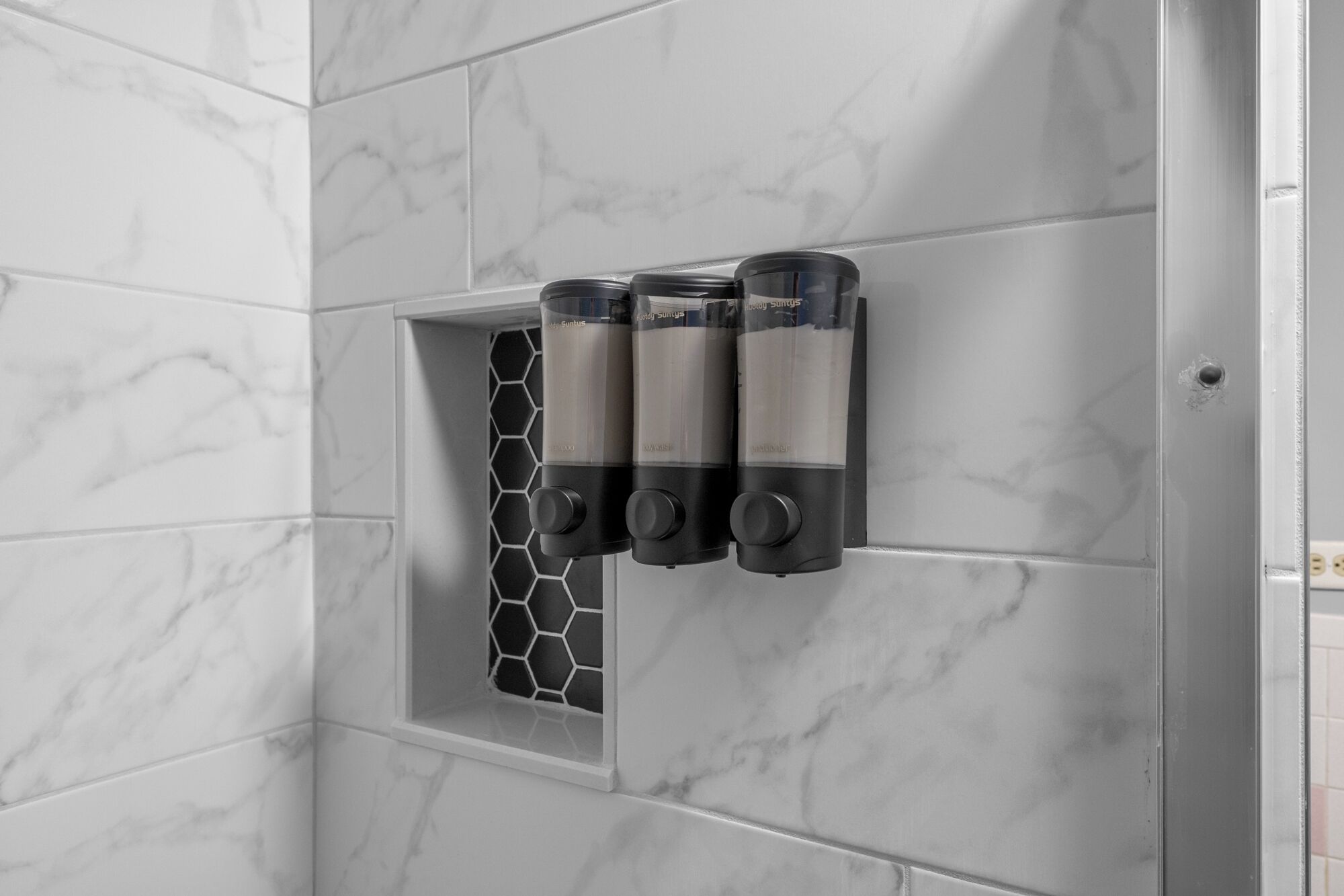 A bathroom with marble walls, a hexagonal window, and three black soap dispensers mounted on the wall.