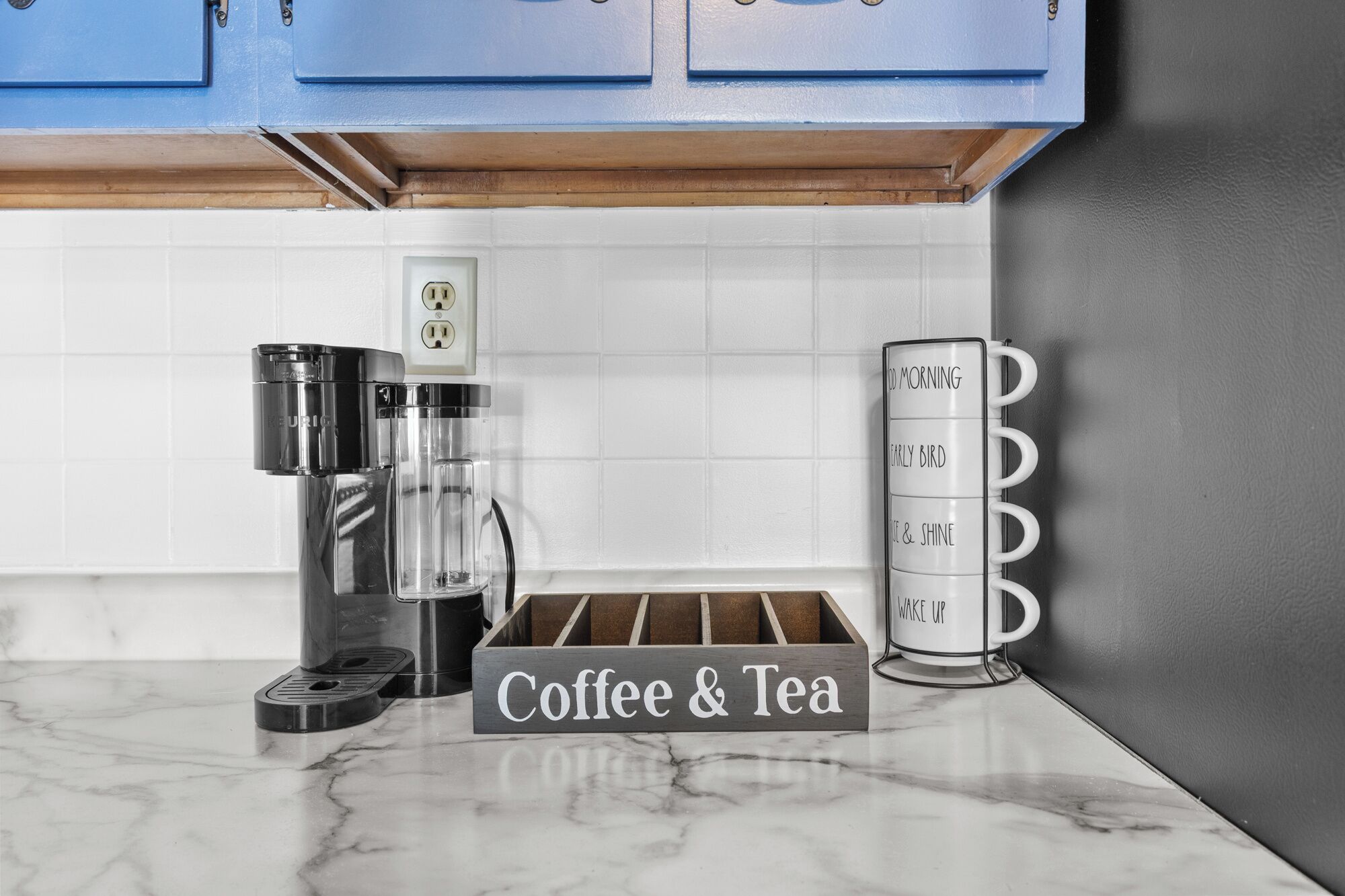 Una cocina con cafetera, cartel de café y té, y un perchero para tazas.