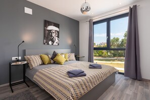 Ein modernes Schlafzimmer mit einem großen Bett, einem Nachttisch, einem Wandbild und Blick durch ein großes Fenster.