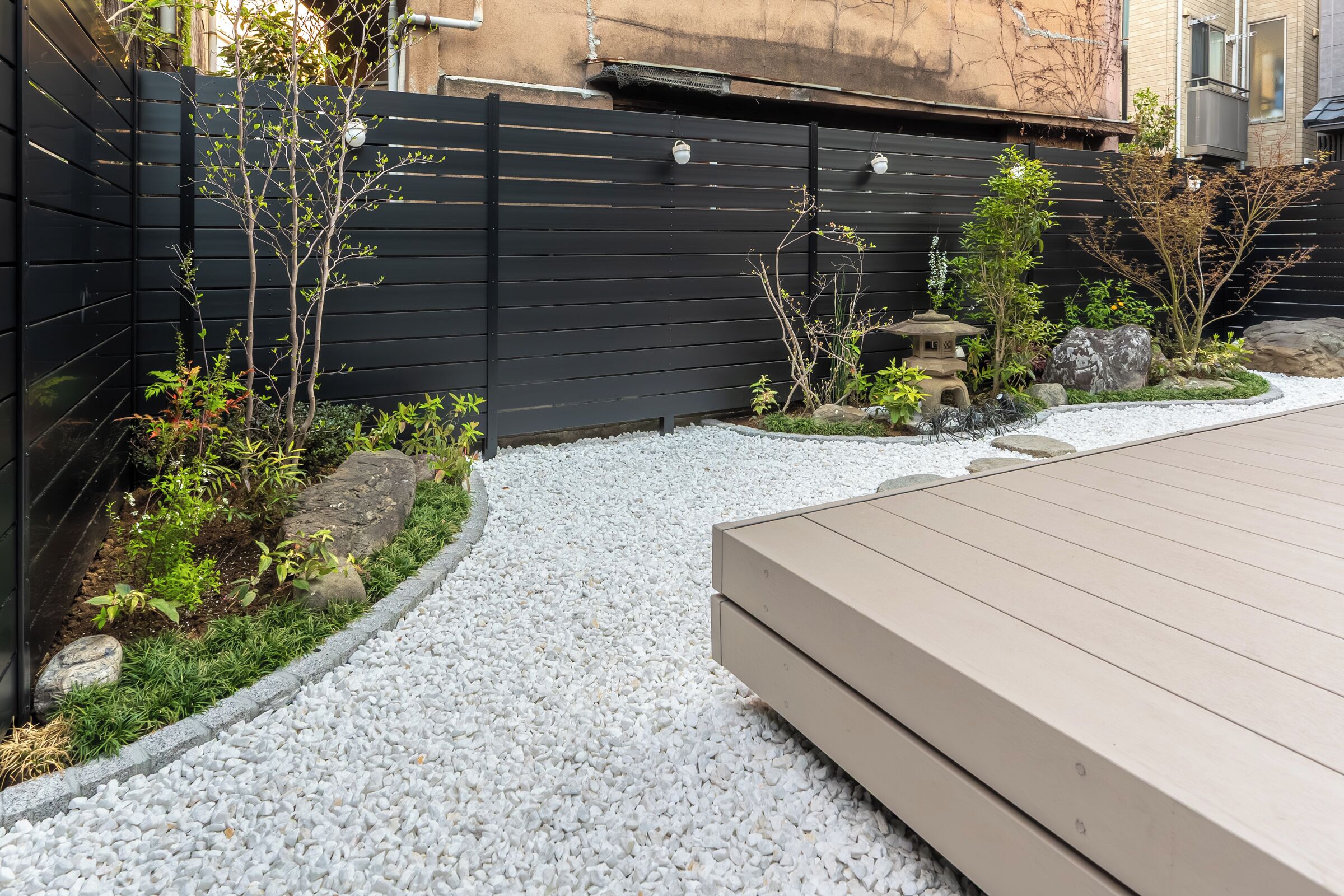 Un jardín con un camino de grava, una terraza de madera y una valla negra.