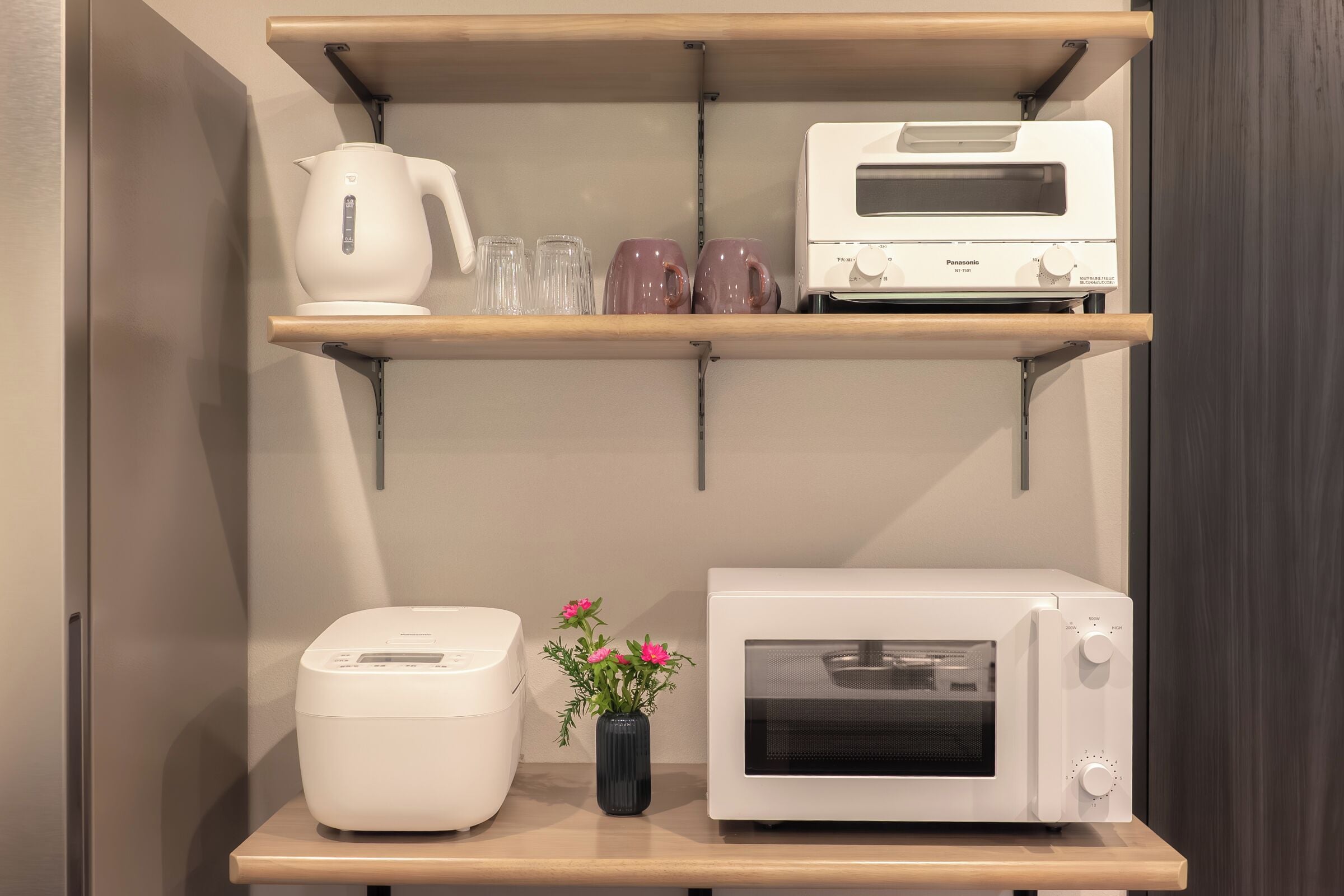 A kitchen with a toaster, a microwave, a kettle, and a shelf with glasses and mugs.