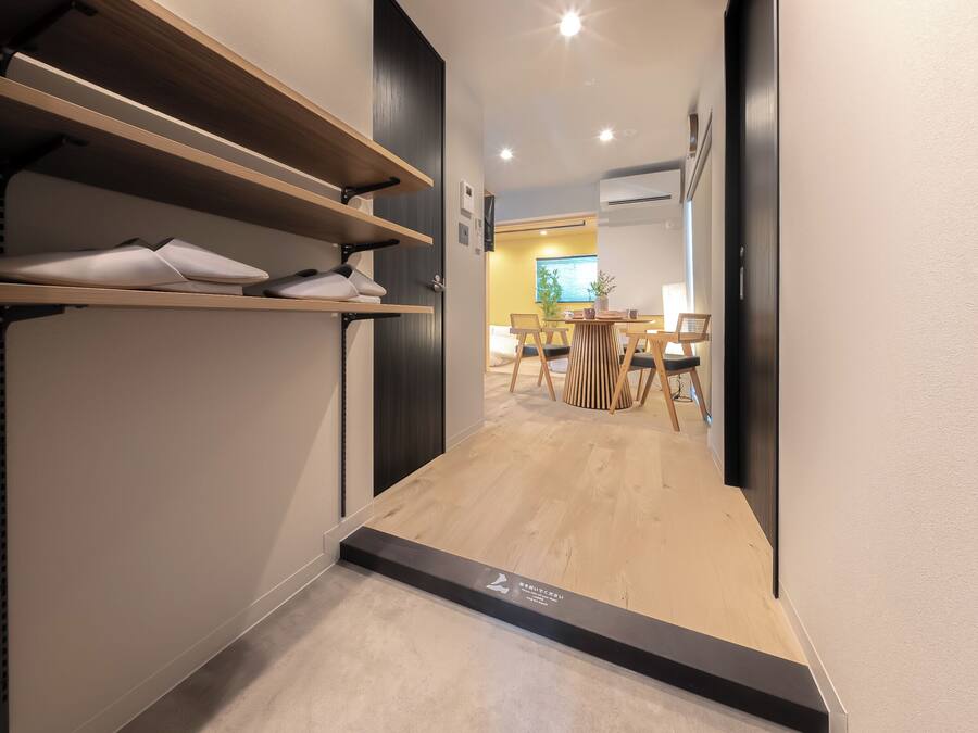 A hallway with wooden flooring, a shoe rack, and a dining area with chairs and a table.