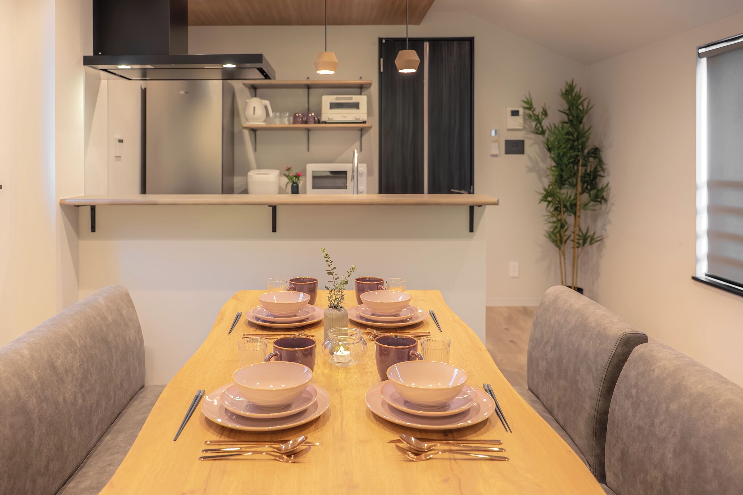 A dining table set with plates, bowls, and cutlery, flanked by sofas, with a kitchen area above and a potted plant to the side.