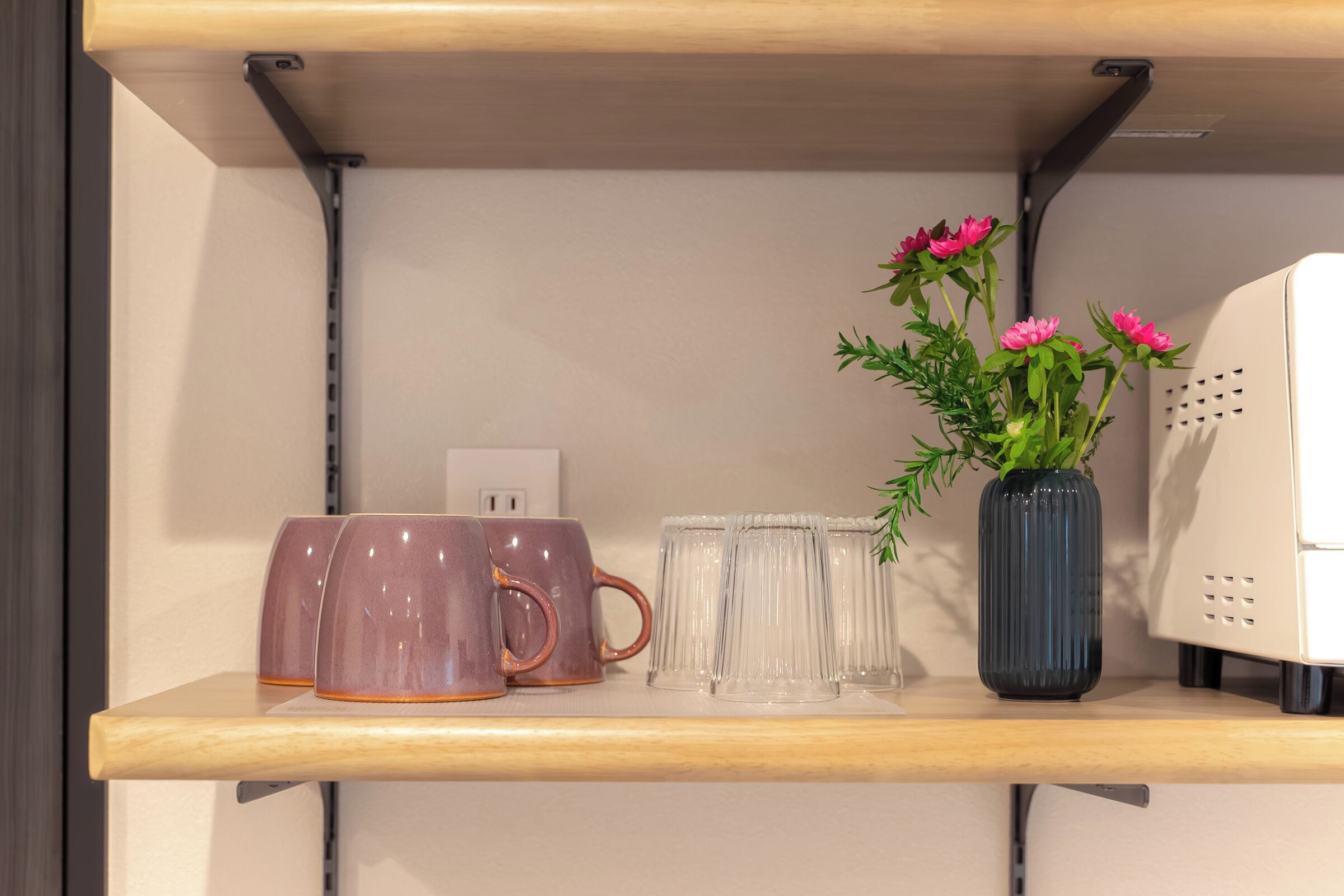 A shelf with ceramic mugs, clear glass tumblers, and a vase with flowers.