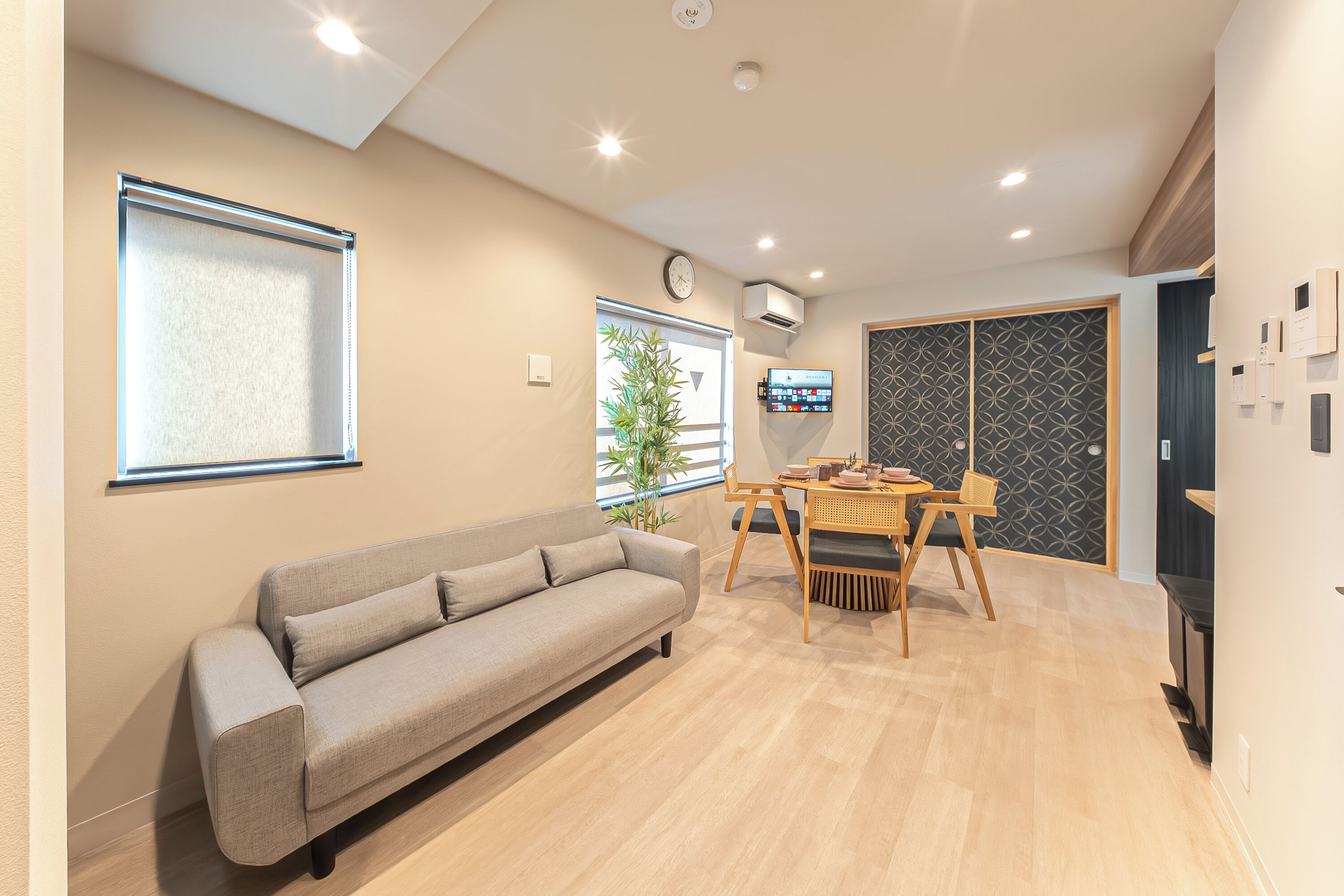 A modern living room with a sofa, a dining table with chairs, a TV mounted on the wall, and a window with blinds.