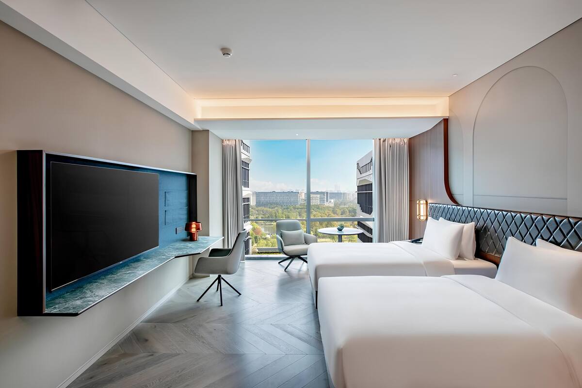 Habitación de hotel con un televisor grande, dos camas, un escritorio y vistas a la ciudad.