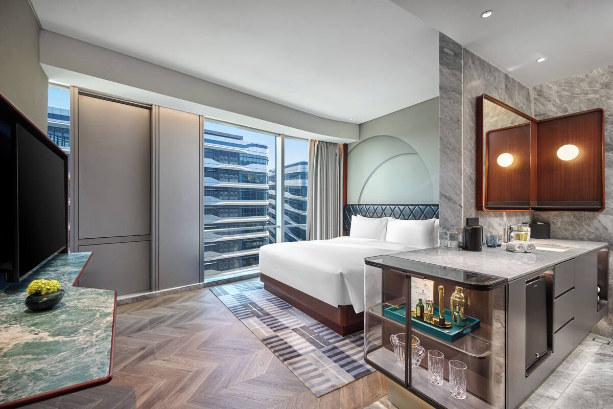 Una habitación de hotel moderna con una cama grande, un tocador con espejo, televisión y vistas a la ciudad.