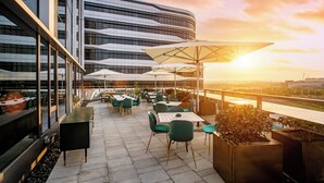 Ein moderner Essbereich im Freien mit Tischen und Stühlen, großen Sonnenschirmen und Blick auf ein Gebäude mit einer geschwungenen Fassade.