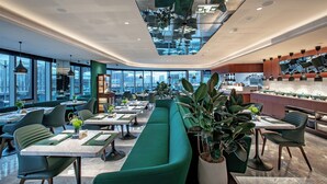 Ein modernes Restaurant mit einem grünen Sofa, Esstischen und Stühlen, großen Fenstern und einer hohen Decke mit hängenden Lampen.