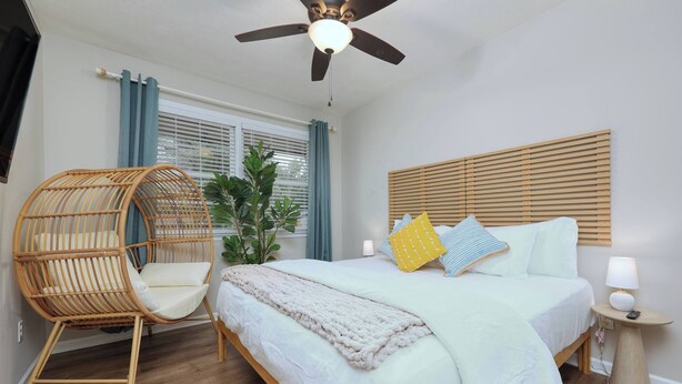 Ein Schlafzimmer mit Bett, Deckenventilator, Nachttisch mit Lampe, Korbsessel und einem Fenster mit Vorhängen.