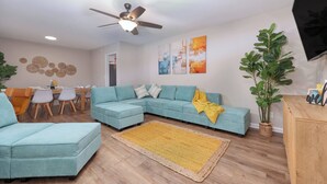 Ein Wohnzimmer mit Ecksofa, Couchtisch, Esstisch mit Stühlen, Deckenventilator und Wandbildern.