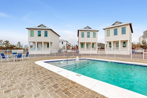 A swimming pool with a brick deck, surrounded by white fences and buildings with a colonial architectural style.