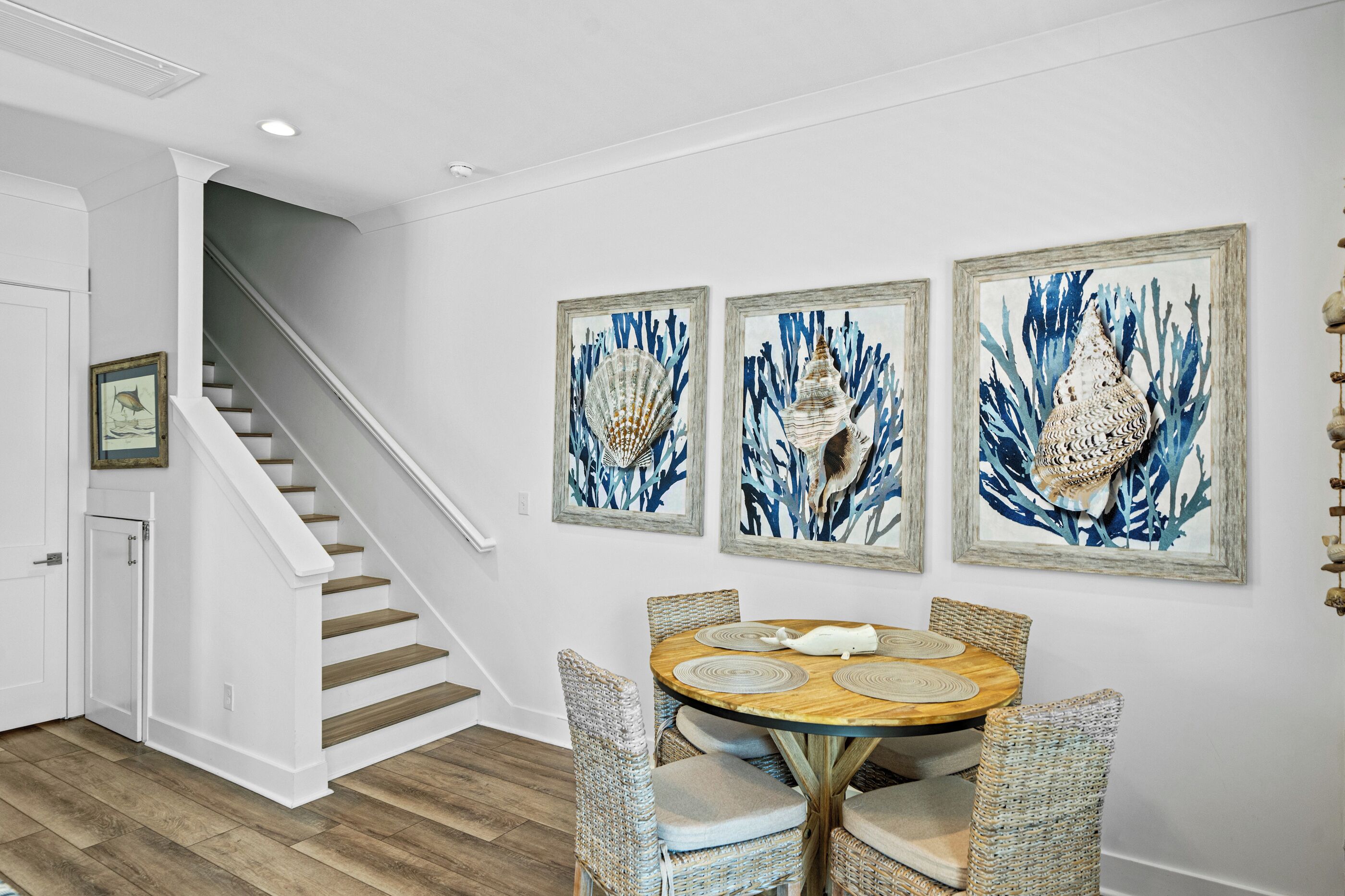A dining area with a round table, four wicker chairs, and three framed artworks on the wall.