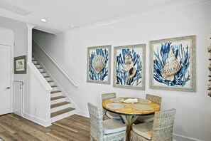 A dining area with a round table, four wicker chairs, and three framed artworks on the wall.