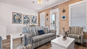 A living room with a striped sofa, a coffee table, and a ceiling fan. There are framed pictures on the wall and a sign that reads 'Orange Beach.'