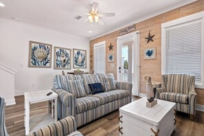 A living room with a striped sofa, a coffee table, and a ceiling fan. There are framed pictures on the wall and a sign that reads 'Orange Beach.'