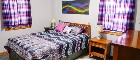 Un dormitorio con cama, mesitas de noche, lámpara, un cuadro y cortinas.