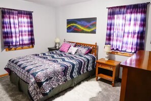 Un dormitorio con cama, mesitas de noche, lámpara, un cuadro y cortinas.