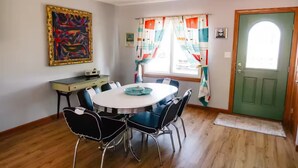 Una zona de menjador amb una taula rodona, cadires, un escriptori, un quadre, una finestra amb cortines i una porta verda.