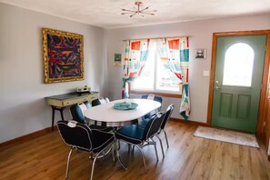 Un comedor con una mesa redonda, sillas, un escritorio, un cuadro, una ventana con cortinas y una puerta verde.