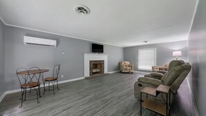 Un salon spacieux aux murs gris, doté d’une cheminée, d’un téléviseur fixé au-dessus, et d’un coin salon comprenant un canapé et des fauteuils.