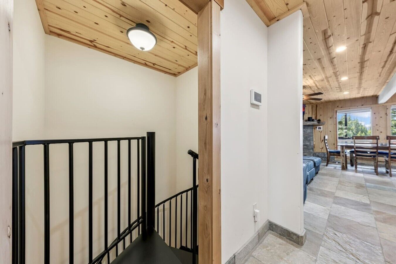 Un escalier en bois avec une rampe noire, un sol carrelé en pierre et une vue sur un espace de vie doté d’une cheminée en pierre et de meubles en bois.