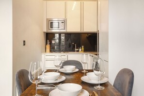 A dining area with a wooden table set for four, a microwave above the counter, and a window with a view. - Chelsea 2BR Flat with Balcony • Fulham Broadway 7min (Greater London)