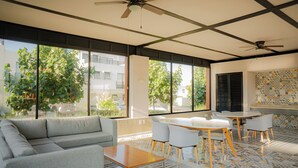 Ein modernes Zimmer mit einer Couch, einem Tisch, Stühlen, einem Deckenventilator, großen Fenstern und einer gefliesten Wand.