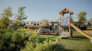 Ein Spielplatz mit Rutsche und Klettergerüst, umgeben von Grünflächen und einem Gebäude im Hintergrund.