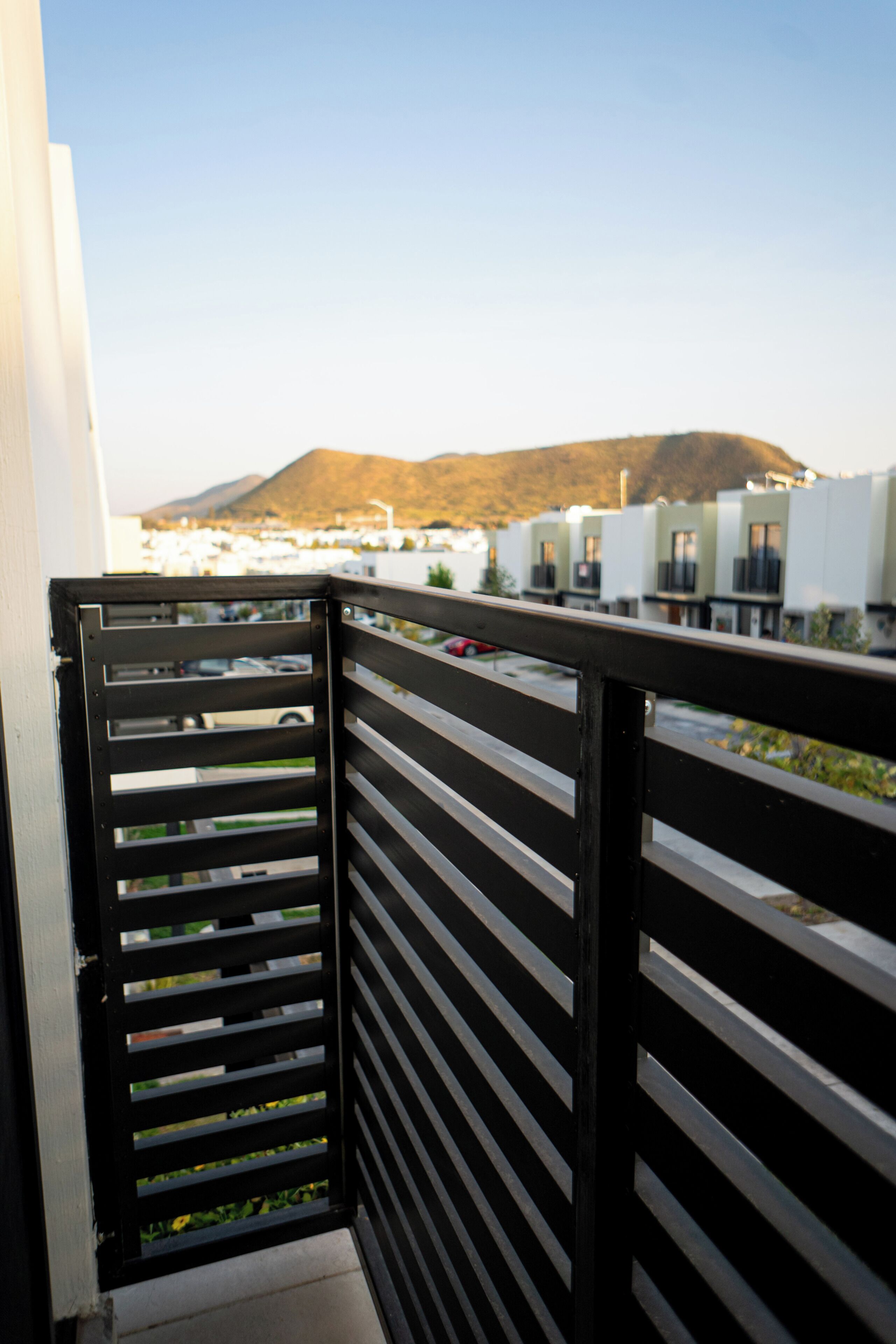 A balcony with a black railing overlooking a parking area and a mountain in the distance.
