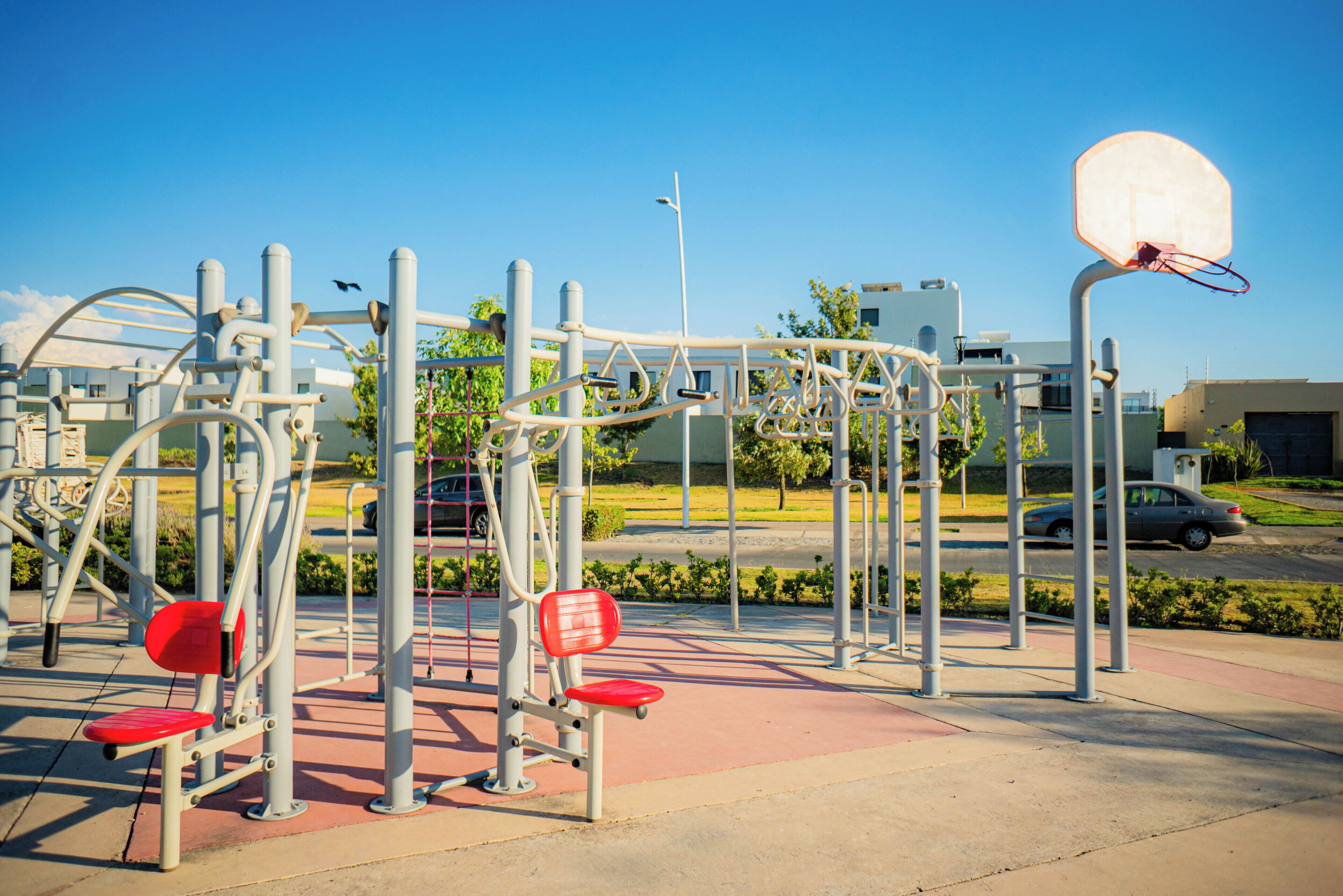 Zona de ejercicios al aire libre con canasta de baloncesto, asientos rojos de gimnasia y estructuras metálicas.