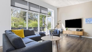 Ein modernes Wohnzimmer mit einem grauen Sofa, einem Couchtisch, einem Fernseher und einem großen Fenster mit Jalousien.