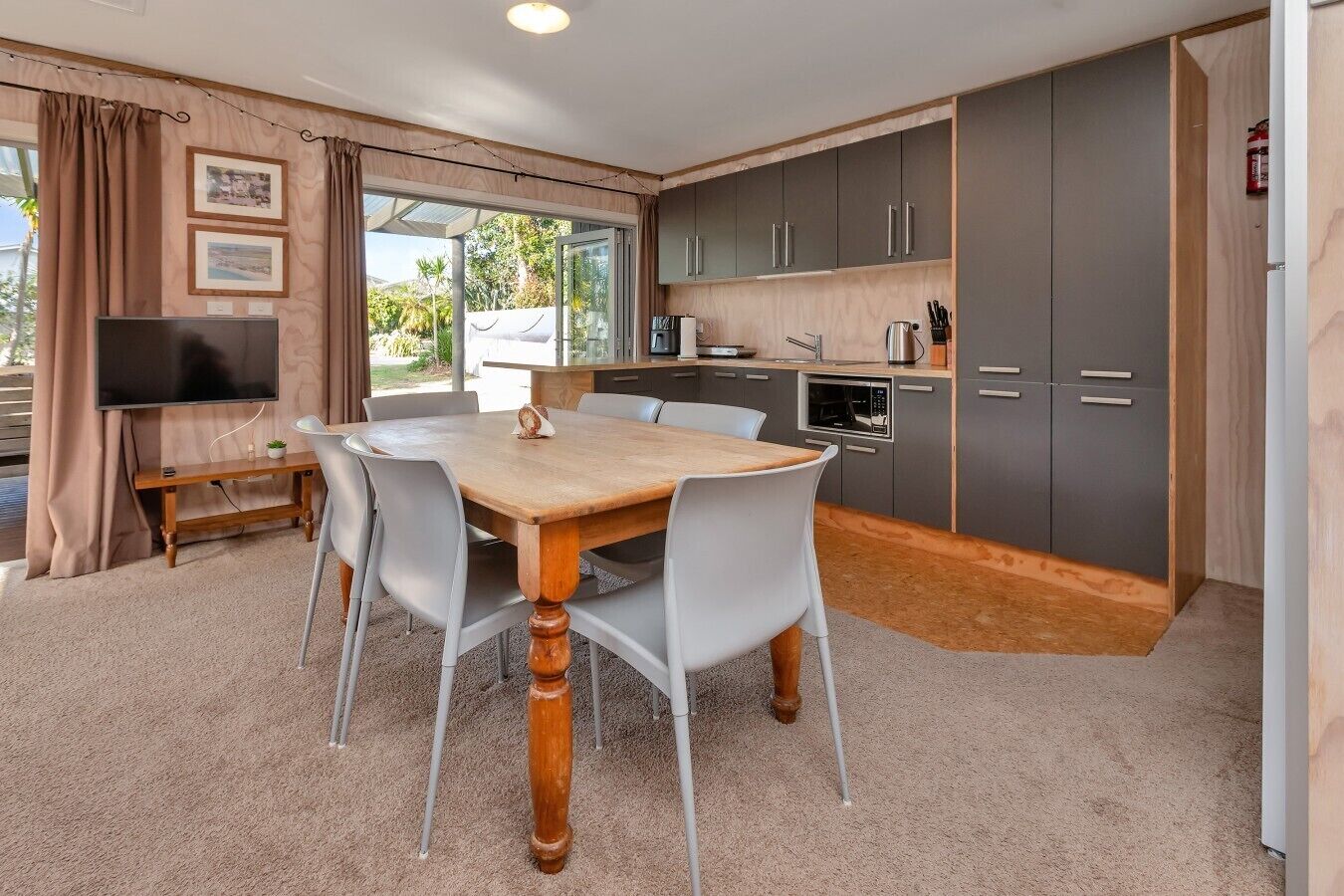A modern kitchen with grey cabinets, a wooden dining table, and a TV in the living area.
