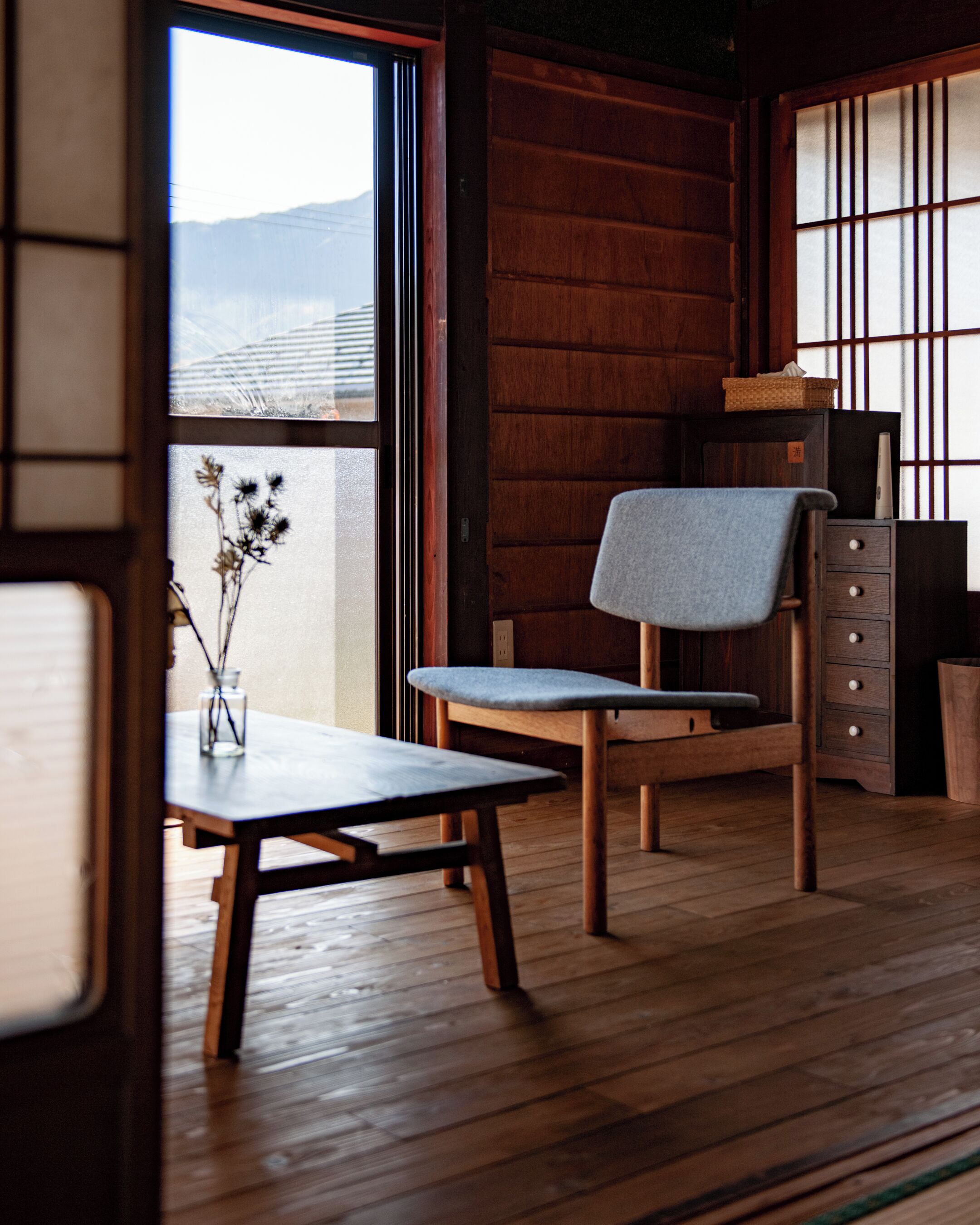 A traditional Japanese room with tatami flooring, a low wooden table, a chair with a grey cushion, and a sliding shoji screen.