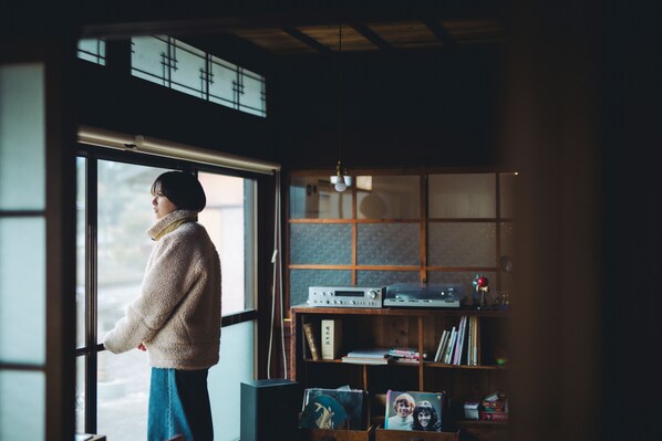 A person standing by a window in a room with a wooden bookshelf and a radio.