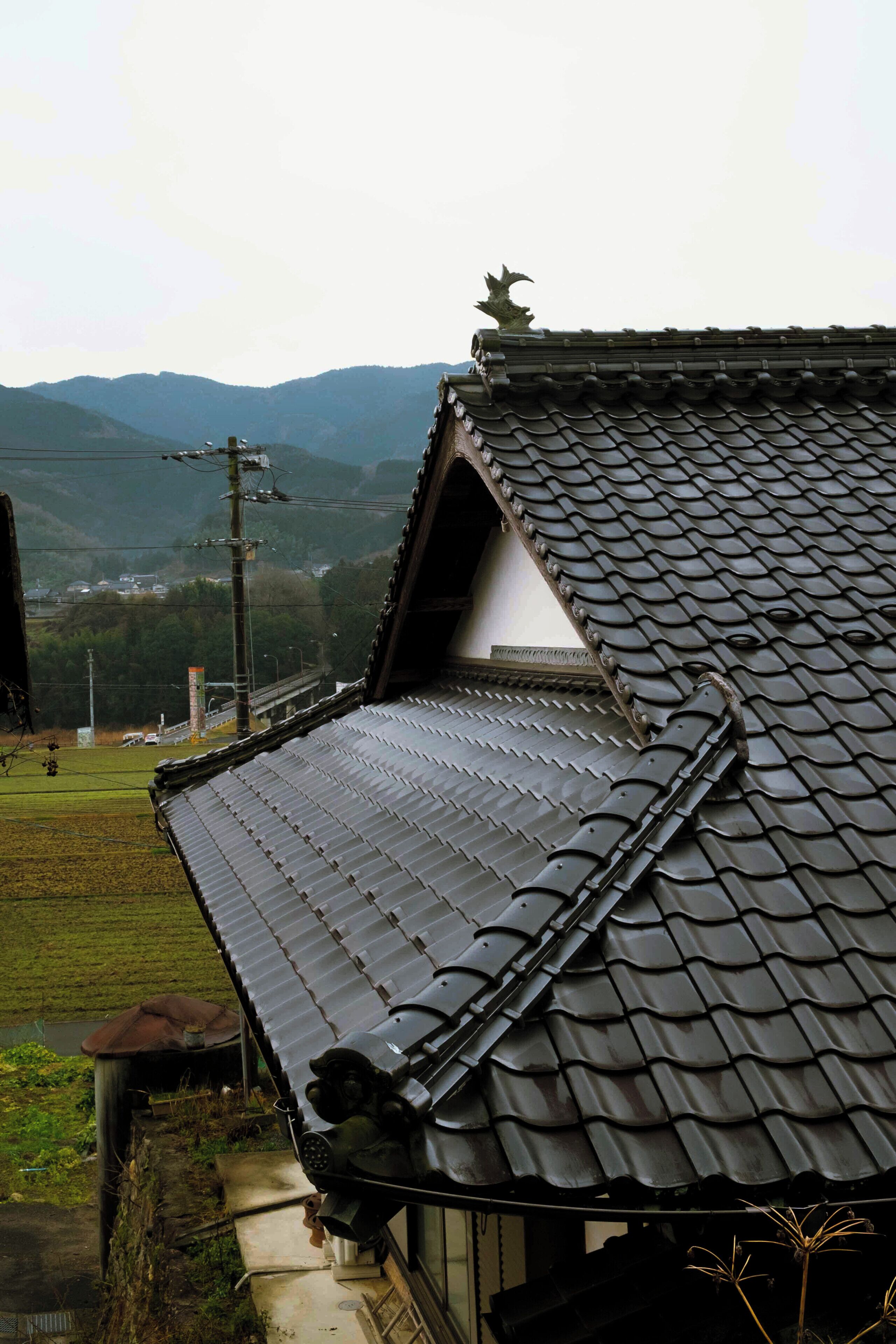A traditional building with a sloped roof, a visible chimney, and a view of a hilly landscape in the background.