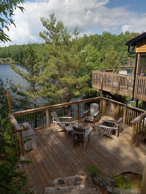 Eine Holzterrasse mit Adirondack-Stühlen und einem kleinen Tisch, mit Blick auf einen See mit Bäumen.